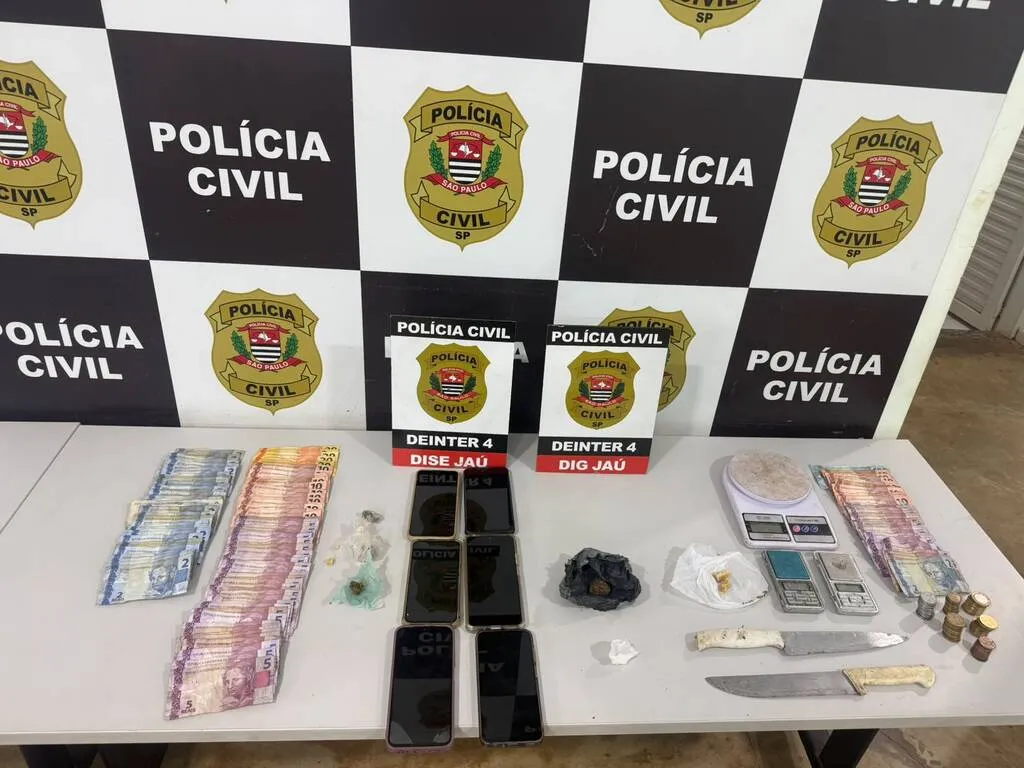 Dinheiro, aparelhos de telefone celular e outros objetos colocados em uma mesa com um painel da Polícia Civil ao fundo