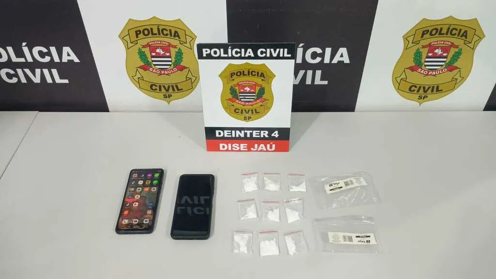 Mesa com dois aparelhos de telefone celular, porções de pós branco com um painel da Polícia Civil ao fundo