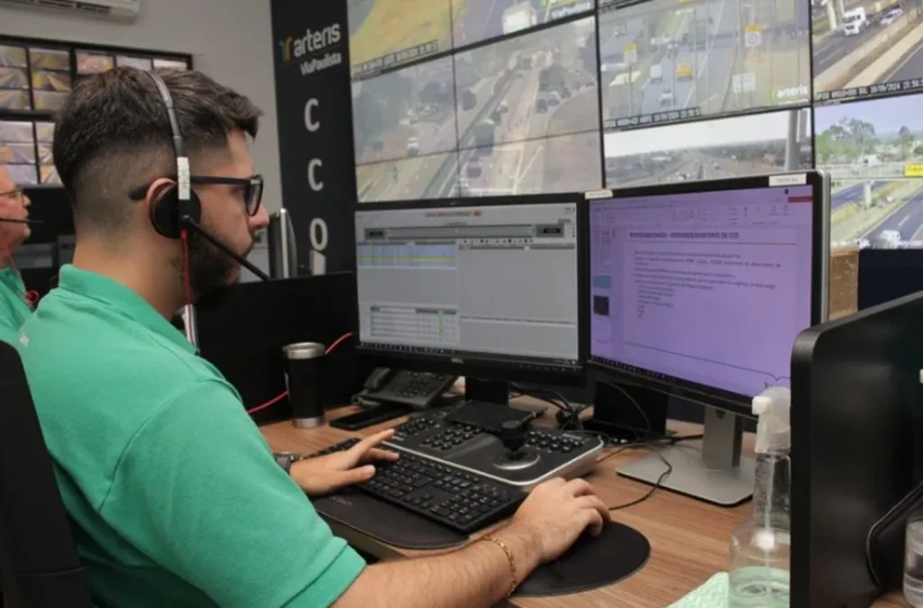 Um operador de camisa verde e headset trabalha em uma estação com dois monitores em uma sala de controle. Ao fundo, um grande videowall exibe imagens de câmeras de monitoramento de tráfego em tempo real.