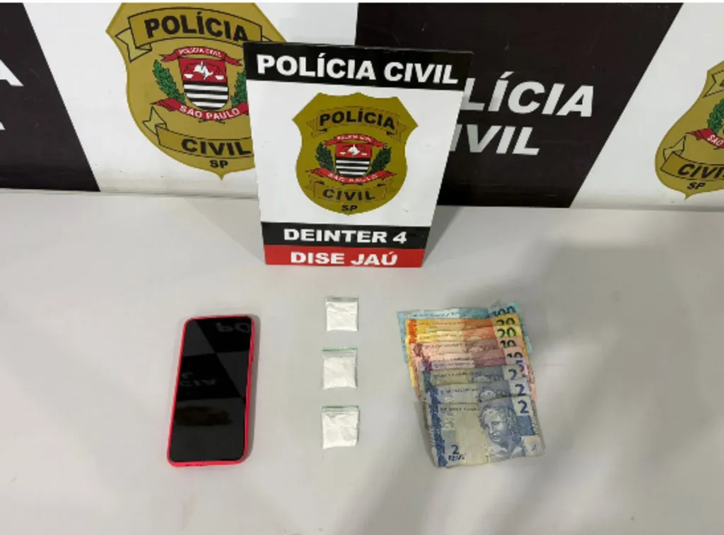 Superfície contendo um telefone celular, três porções de pó na cor branca e dinheiro em cédulas diversas, além de um painel ao fundo com o símbolo da Polícia Civil do Estado de São Paulo