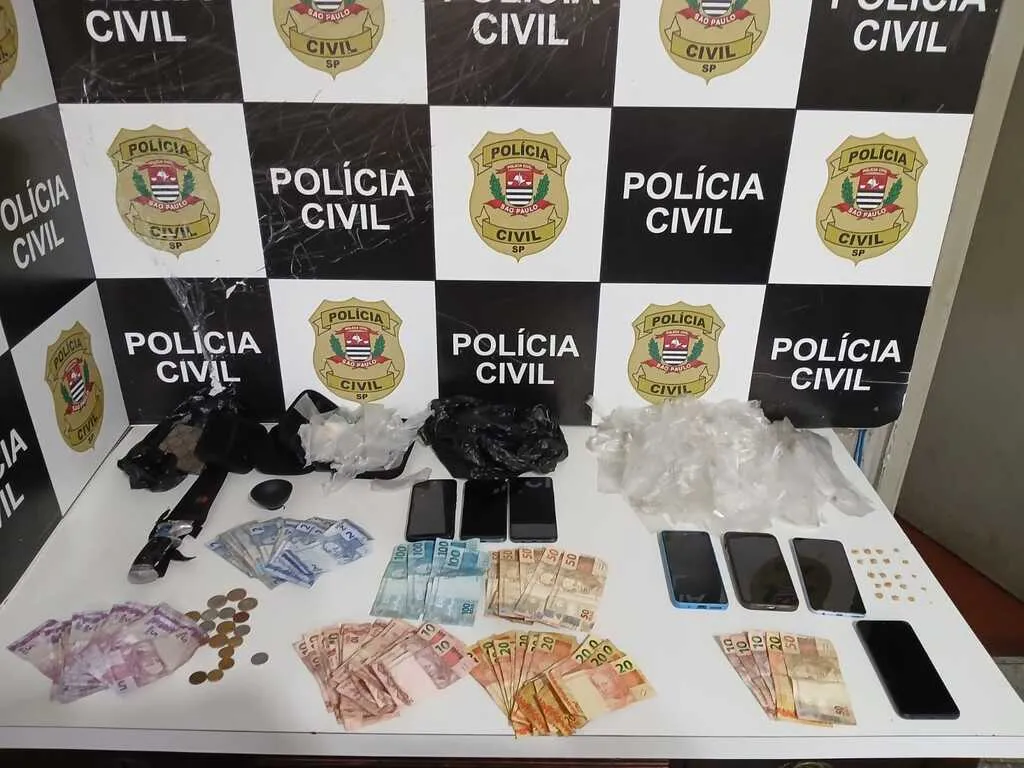 Mesa na cor branca com dinheiro, celulares e sacos plásticos, tendo painel ao fundo escrito Polícia Civil