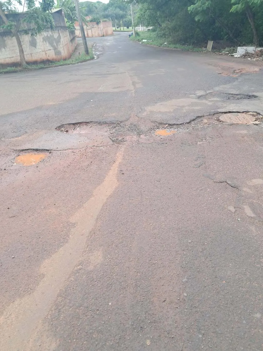 Rua asfaltada, com buracos e muro do lado esquerdo e ao fundo, tendo vegetação no lado direito