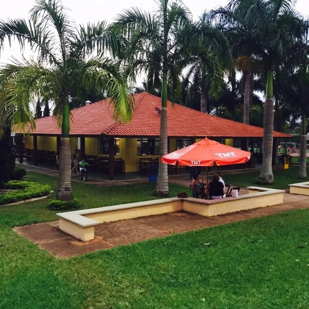 Construção com paredes amarelas e telhado marrom, com vegetação verde em volta além de mesa com pessoas em um guarda sol vermelho