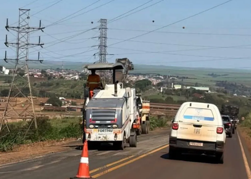 Maquinário de pavimentação asfáltica e veículo de apoio realizando manutenção em uma rodovia movimentada.