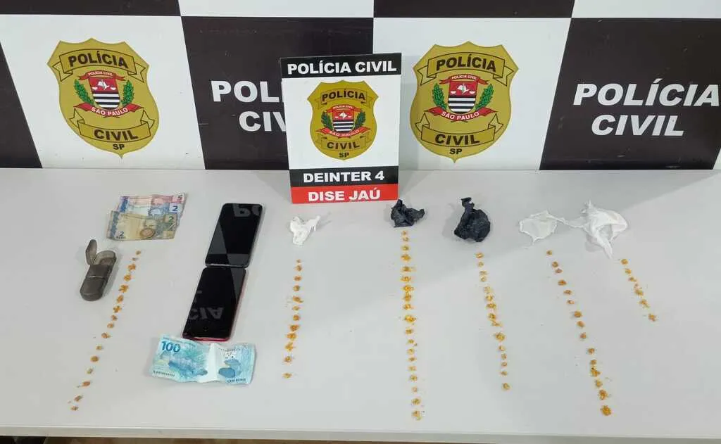 Mesa na cor branca contendo dinheiro, celulares, porções de drogas na cor amarela e painel da Polícia Civil ao fundo