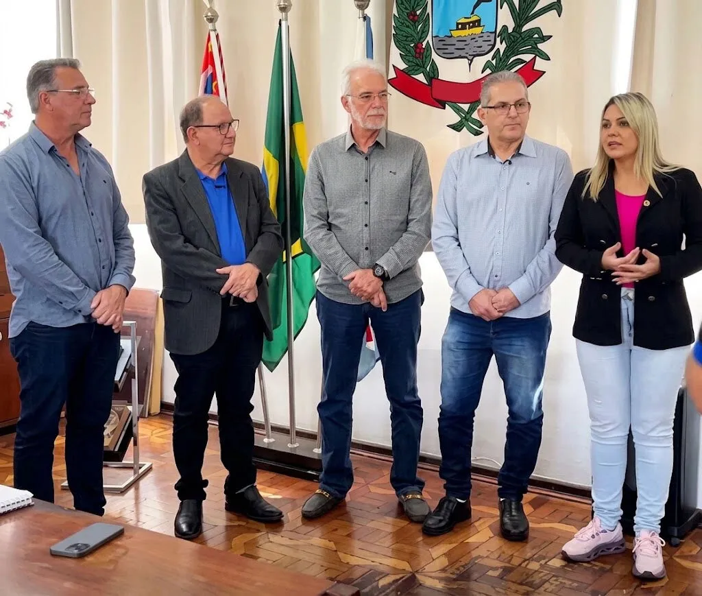 quatro homens e uma mulher em pé numa sala com piso de madeira, bandeiras e brasão ao fundo
