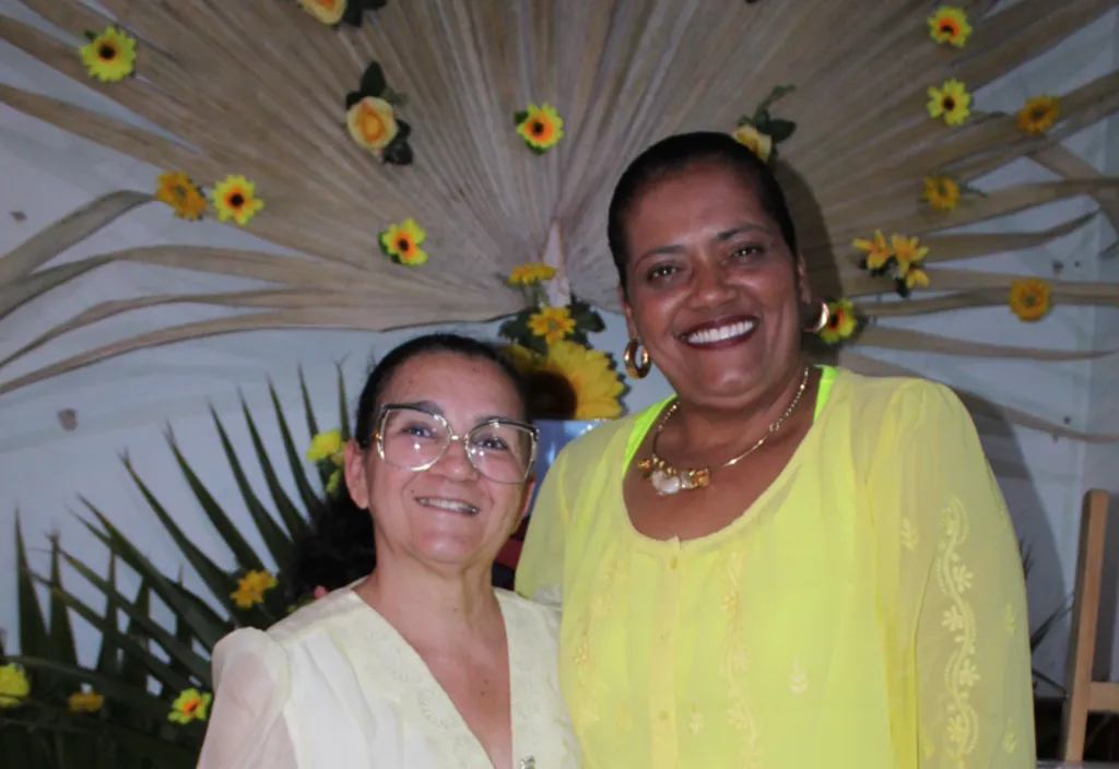 duas mulheres de amarelo posam para foto, uma branca idosa e uma negra de meia idade