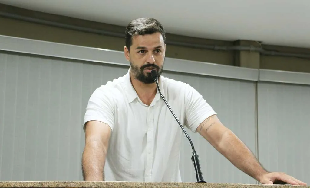 Homem de barca e camisa social branca, apoiado sobre uma mesa fala ao microfone