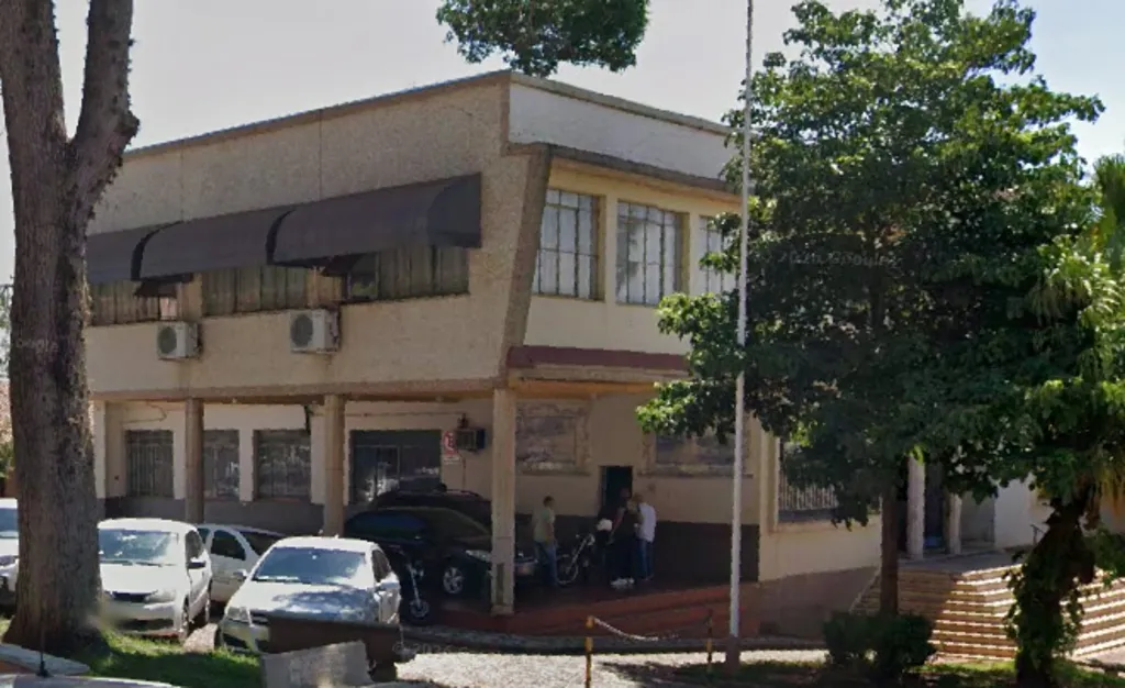 Prédio de dois andares, cercado por árvores, escada do lado direito, veículos estacionados e pessoas no térreo