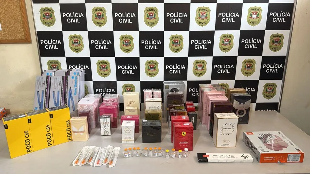 Mesa com embalagens diversas e ampolas de medicamentos, tendo ao fundo painel da Polícia Civil