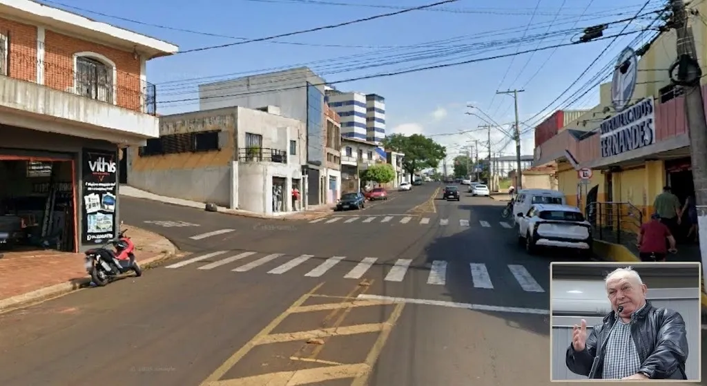 Via pública com faixa de pedestres e veículos estacionados dos dois lados da rua, tendo um homem de blusa preta com a mão levantada no detalhe