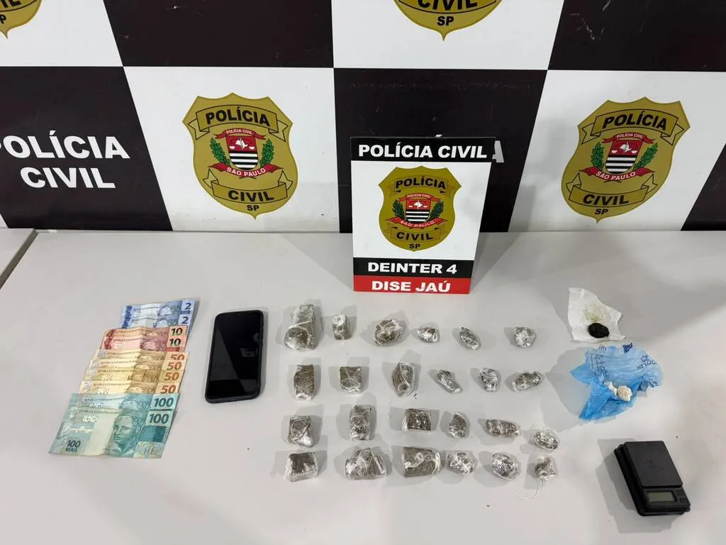 Mesa na cor branca contendo dinheiro, celular, porções de maconha embaladas em plástico, com painel da Polícia Civil ao fundo