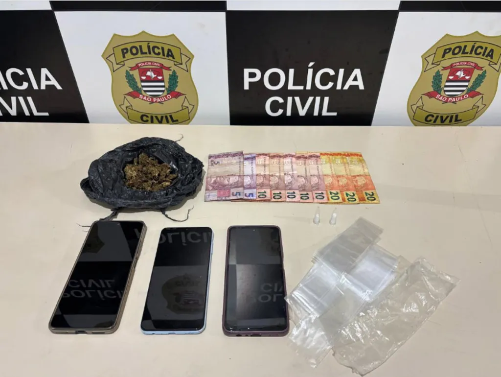 Telefones celulares, embalagens brancas, dinheiro e saco preto contendo maconha, com um painel da Policia Civil ao fundo