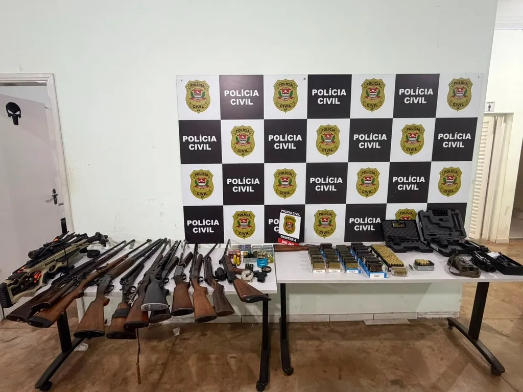 Mesas com diversas armas, munições e artigos relacionados, tendo atrás um painel escrito Polícia Civil
