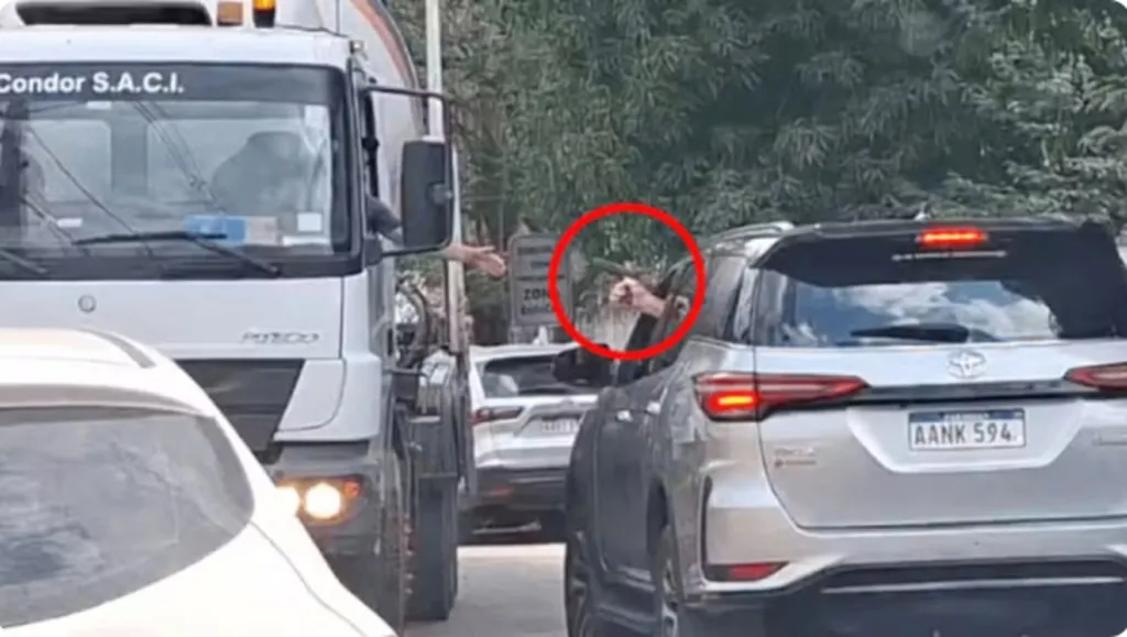 Veículos transitando em uma rua, onde o condutor de um carro aponta arma para o motorista de um caminhão que vem em sentido contrário