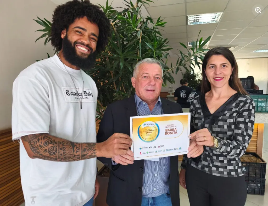Três pessoas sorriem em um ambiente interno enquanto seguram juntas o certificado de transparência da Câmara Municipal de Barra Bonita.