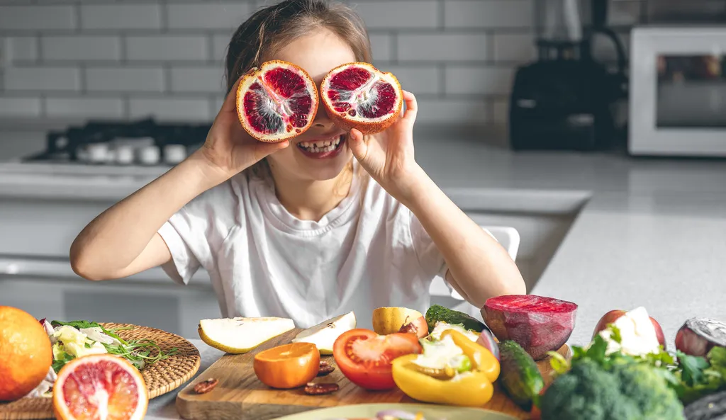 criança sentada à mesa brinca com rodelas de laranja em frente aos olhos, como um óculos, com diverss legumes à frente